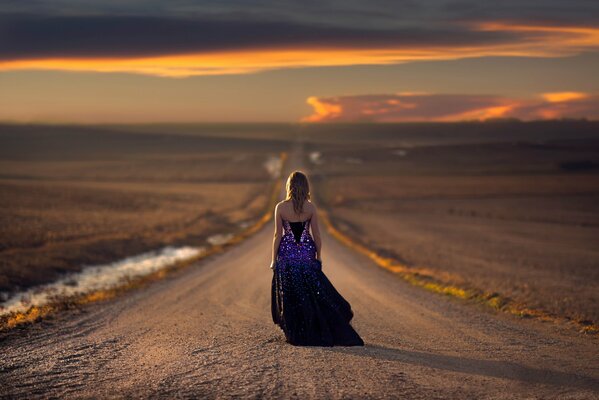 A girl walks down the road into the sunset in an evening dress