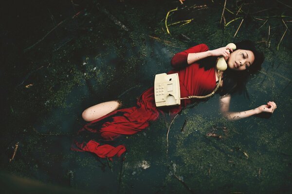 A girl in a red dress with a phone is lying in a swamp