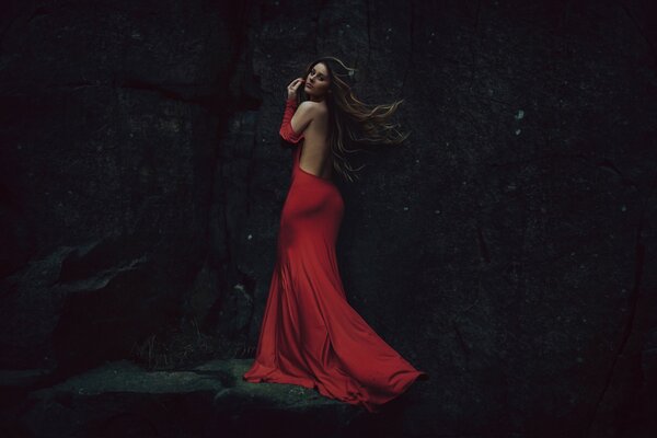 A beautiful girl in a red dress with an open back against a dark rock background