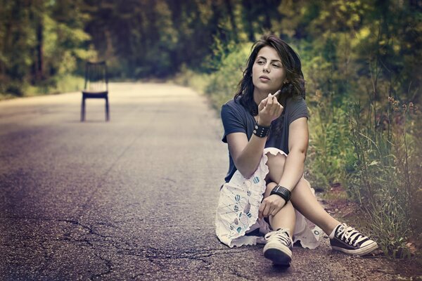 The girl is sitting on the side of the road, on the ground and smoking, there is a chair in the middle of the road