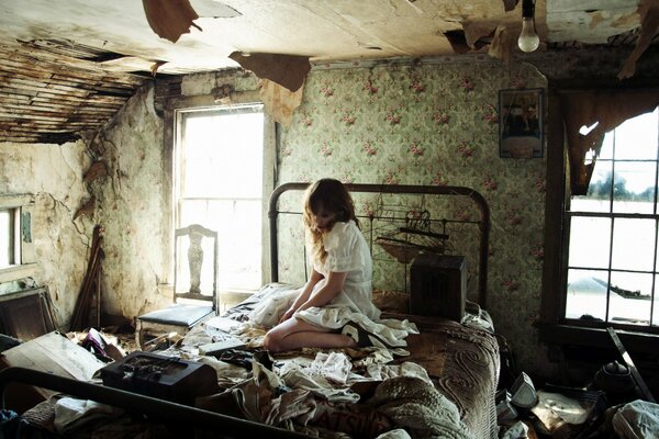 A girl in a white dress is sitting on a cot among scattered things