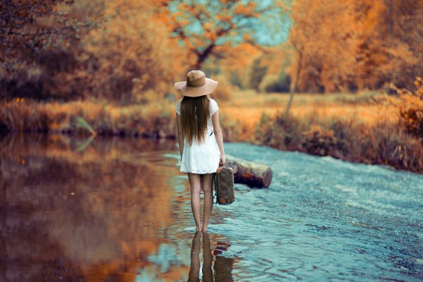 Ragazza in abito corto bianco e cappello cammina lungo il fiume con una valigia, vista posteriore