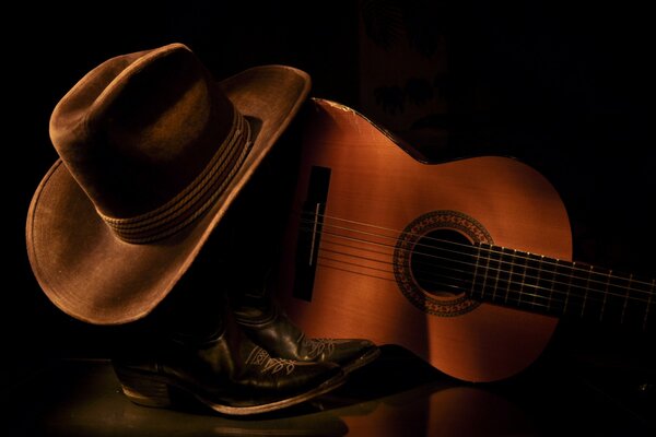 Cowboy boots and a hat are lying near the guitar on a dark background