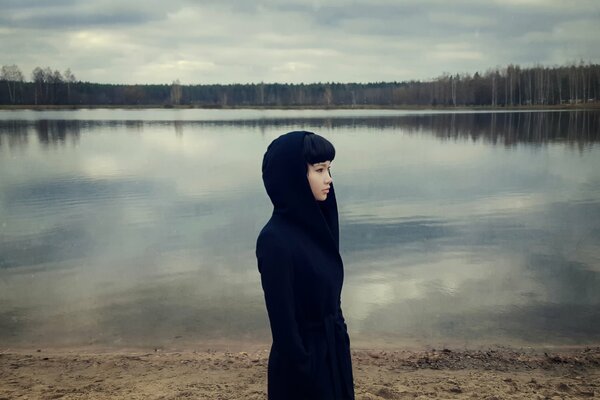 Dramatic photo of a girl in a black coat against the background of a lake and a gray forest without foliage