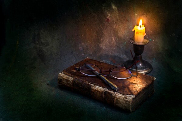 A candle illuminates an old book and glasses