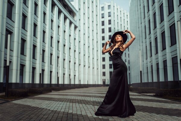 A girl with a beautiful figure in a black dress with a neckline and a hat on the background of the city