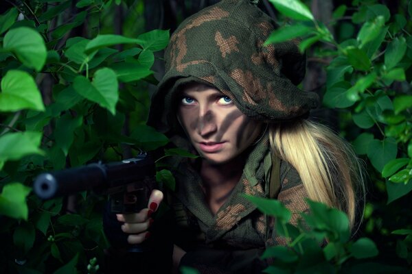 A girl in camouflage clothes and war paint aims a pistol from behind the bushes