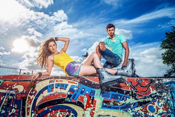 A girl on roller skates lies on a graffiti wall with a guy