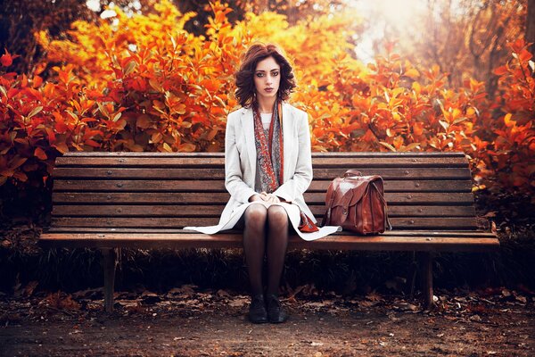 Bruna in un mantello bianco seduto su una panchina nel parco autunnale