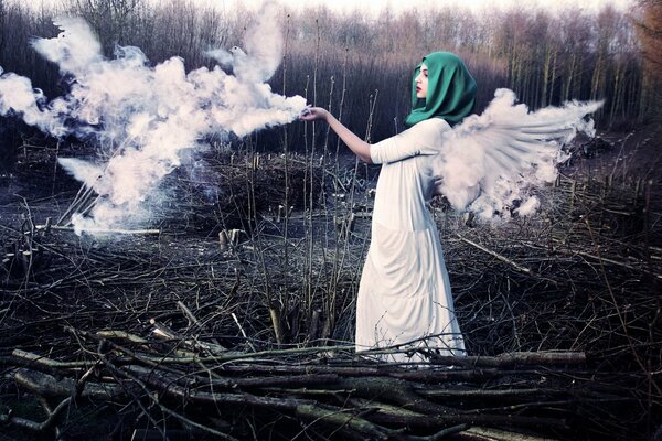 A girl in a simple white dress with wings and a green scarf is standing in a clearing with brushwood