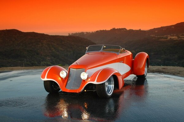 Orange hot rod stands on wet asphalt against the background of hills and sunset