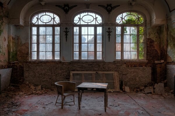 A table and an armchair in a room with three windows in an abandoned building