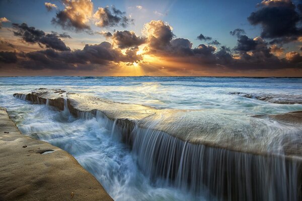 The sun behind the clouds, the sea and the waterfall