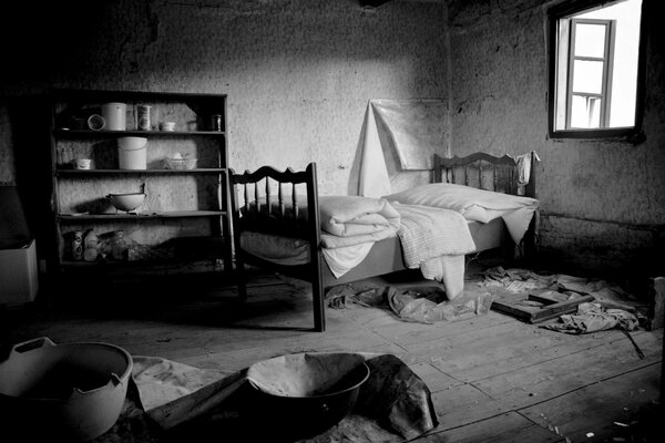 Брошенный людьми дом с покареженными окнами с незаправленной кроватью и полками с оставшейся посудой в черно-белом виде