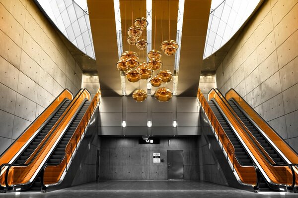 Orange escalators and an orange chandelier in the form of flowers in a room lined with large gray tiles
