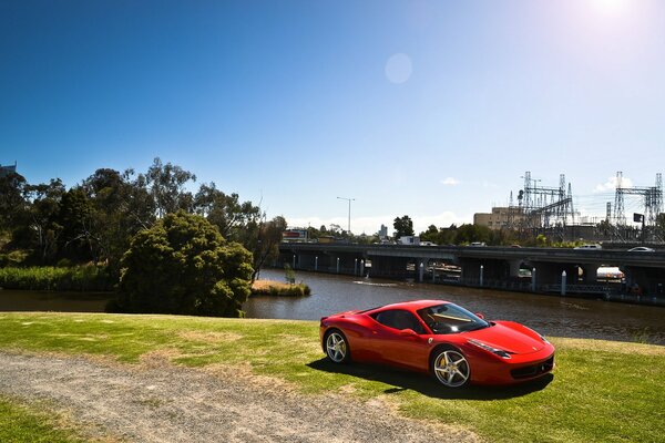 A red Ferrari is standing on the grass near the river