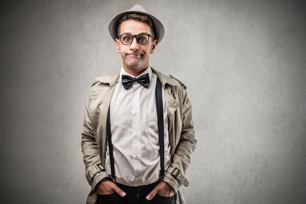 Un hombre con tirantes, sombrero, gafas y abrigo está de pie cerca de una pared gris