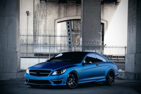 A blue tinted Mercedes-Benz stands between gray columns in front of the fence