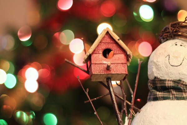 A snowman holds a birdhouse against the background of lights