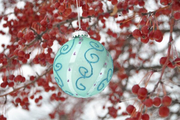 Una bola de año nuevo turquesa con patrones cuelga de hilos blancos contra ramas con pequeñas manzanas rojas en invierno