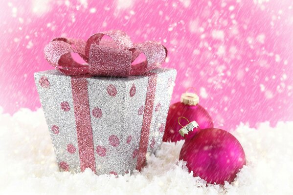 Auf dem Schnee eine Kiste mit einem rosa Band, nebeneinander zwei rosa Weihnachtskugeln und fallendem Schnee