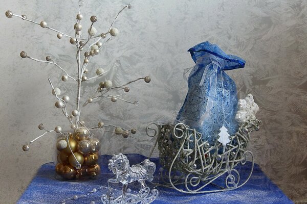 Cavallo di vetro, una borsa blu legata con un nastro in una slitta, perline bianche, installazione di un albero di Natale in un barattolo con palline dorate di Capodanno su una tovaglia blu e bianca contro un muro grigio