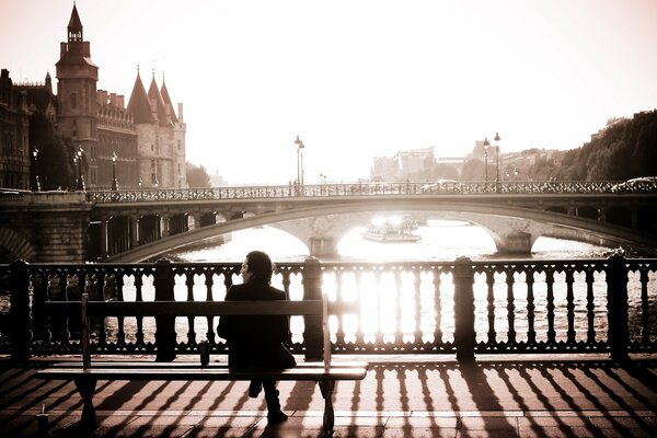 Черно-белое фото . Париж, человек сидящий на мосту на лавочке утром на фоне моста , домов