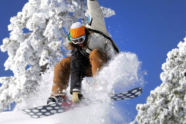 Eine Snowboarderin mit Brille und Mütze fährt unter schneebedeckten Bäumen Snowboarden und hebt einen Haufen Schnee nach oben