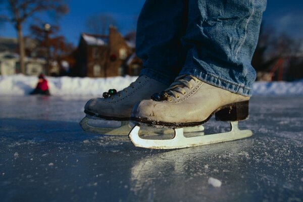 A man in skates and jeans on a skating rink near the house. In the background, a man in a red jacket is sitting on the ice