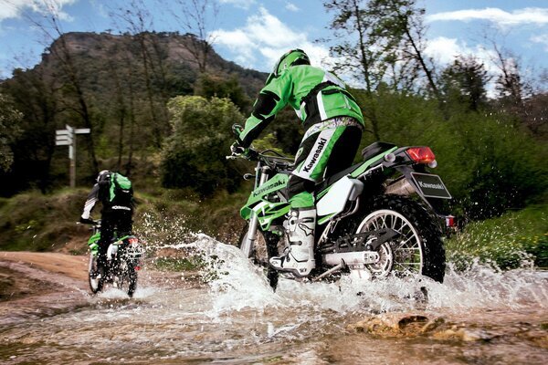 Two motorcyclists in black and green cross a mountain stream