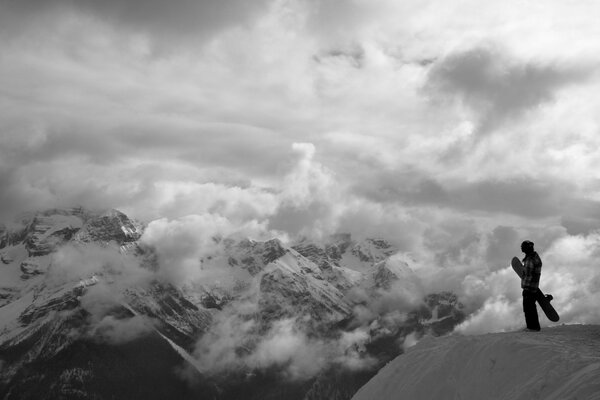 A man with a snowboard in his hands in front of snow-capped mountains, engulfed in clouds. Black and white mood