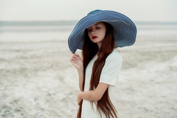 Une fille dans une robe blanche tient un chapeau à larges bords rayé