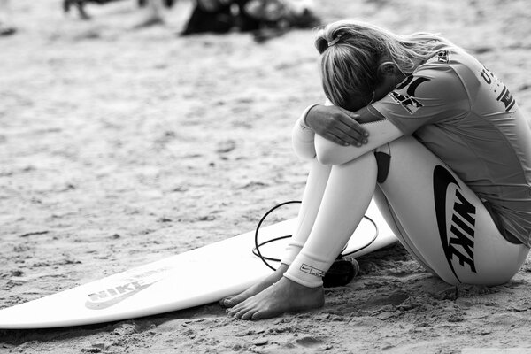 Foto en blanco y negro de una niña enterrando su cabeza en sus rodillas con una tabla de surf tumbada en la arena