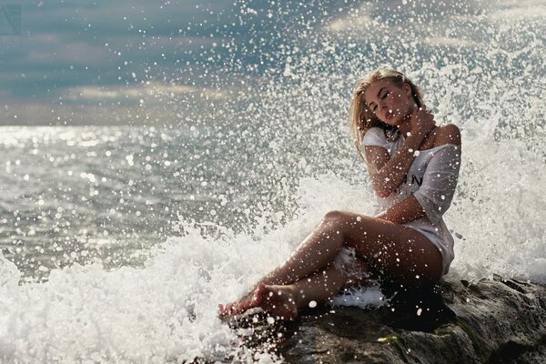 Jeune blonde en vêtements blancs est assis sur le bord de la mer et les éclaboussures