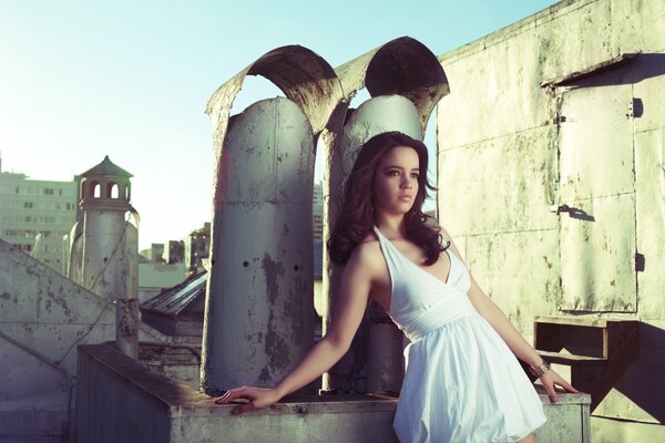 A girl in a white dress against the background of pipes and the roof of a building