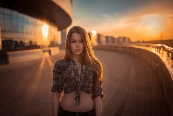 Against the background of the sunset, a girl with loose long hair in a plaid shirt tied above the waist