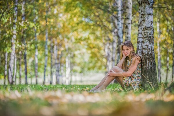 A girl in a dress sitting by a tree in the forest