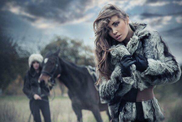 A girl in a fur coat poses against the background of a horse