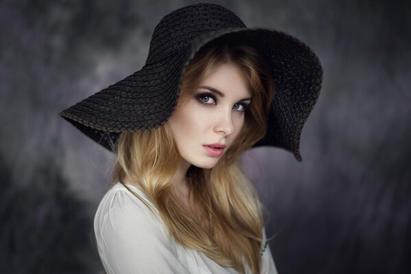 A girl with loose hair and a white blouse in a big black hat on a black background