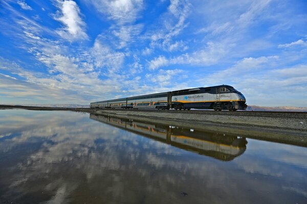 The train travels along the lake, in which clouds are reflected