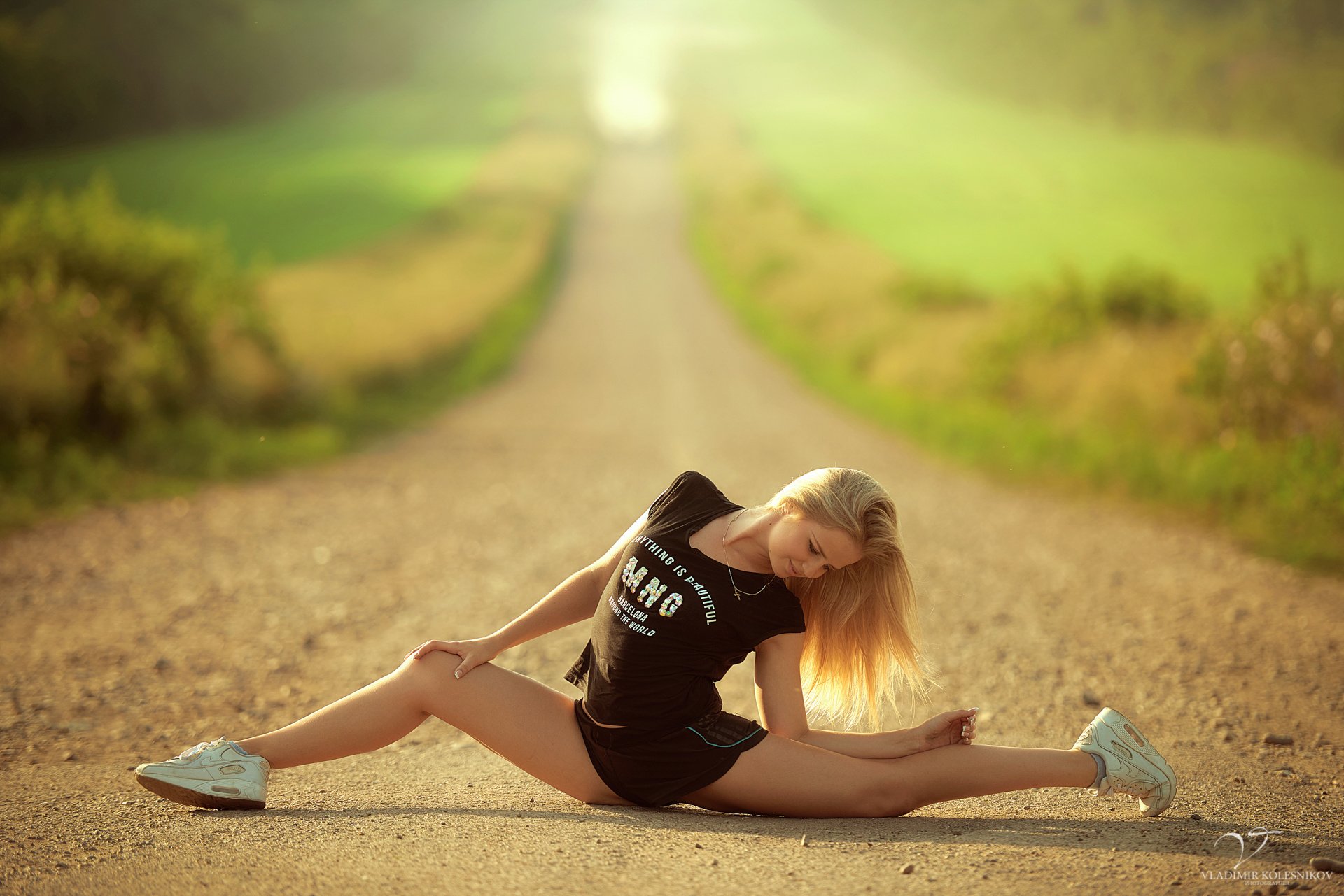 delgado atlético chica rubia pueblo medio carretera pose estiramiento cordel flexibilidad camiseta pantalones cortos zapatillas de deporte sol verde desenfoque