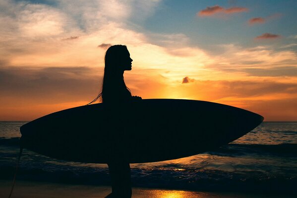 Silhouette eines Mädchens mit einem Surfbrett auf dem Hintergrund des Meeres und eines luxuriösen Sonnenuntergangs