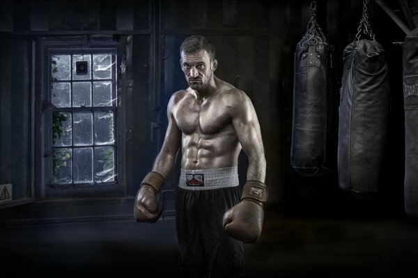 A beefy man with boxing gloves on the background of a window and punching bags