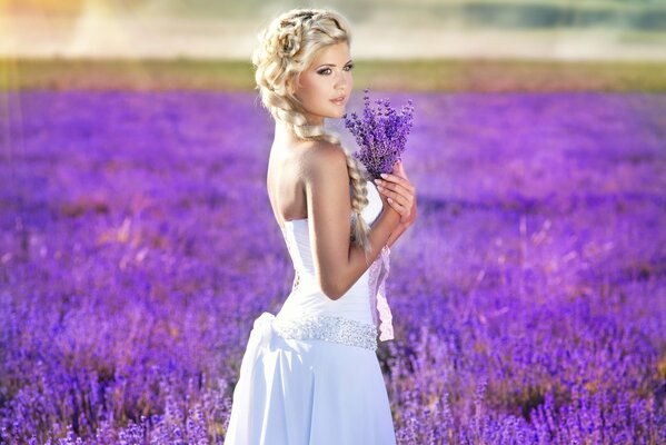 Piękna dziewczyna w białej sukni stoi w polu z bukietem lawendy w dłoniach