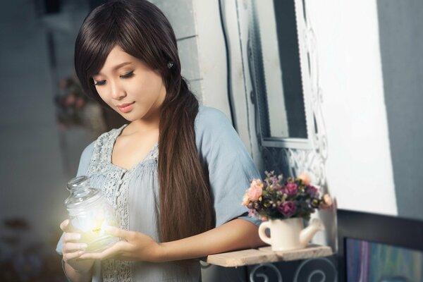 A beautiful Asian woman stands at a vase with flowers and gently holds a decanter