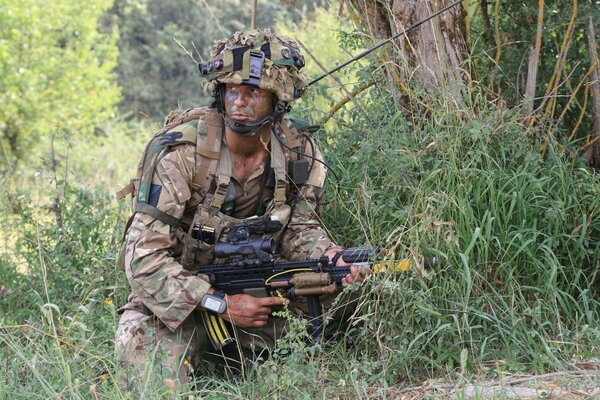 A soldier in camouflage hides in the bushes with a weapon