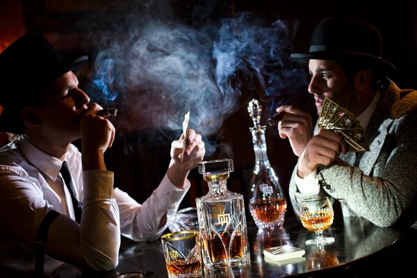 Dwóch mężczyzn gra w karty, pali i pije przy stole