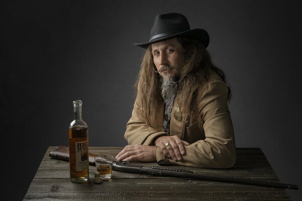 Un homme aux cheveux longs avec une barbe dans un chapeau à l image d un cow-Boy est assis à une table en bois sur laquelle se trouve une bouteille de whisky et un verre à moitié rempli, ainsi qu un fusil à pompe