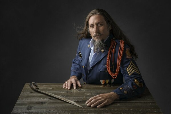 A long-haired man with a beard in a military uniform is sitting at a wooden table on which a combat saber is lying