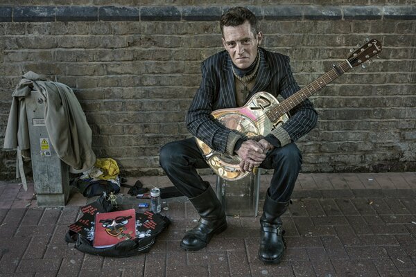 A man with a guitar at a brick wall